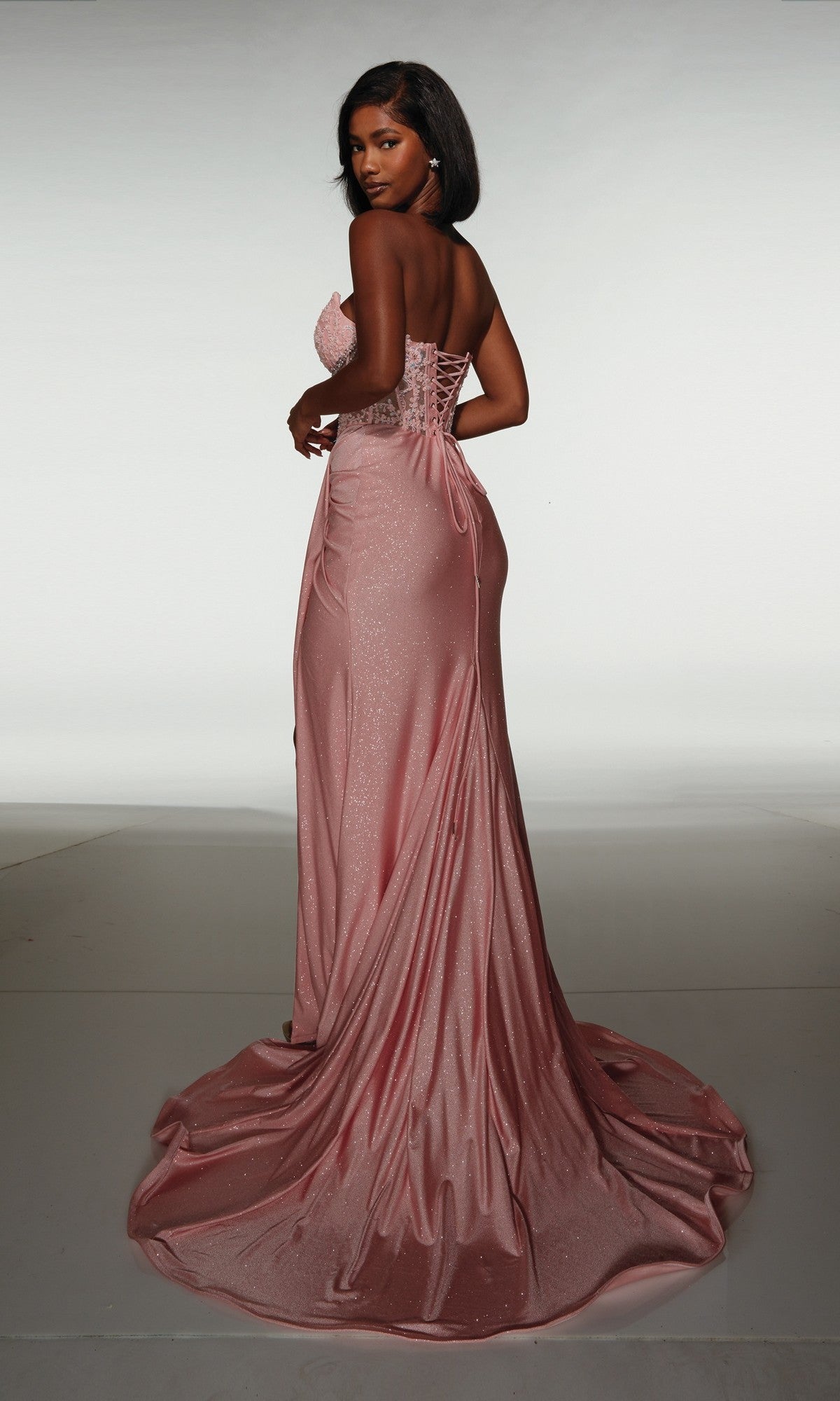 A woman stands with her back to the camera, wearing the Alyce 62057 long formal dress—a strapless, floor-length pink gown with a lace-up back—set against a plain background.